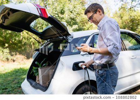 Man with shopping bag next to a charging electric car in the yard of a country house Man with shopping bag next to a charging electric car in the yard of a country house 108802802