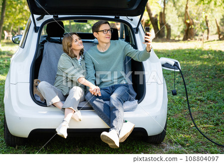 Loving couple with smartphones sits in an electric car's trunk Loving couple with smartphones sits in an electric car's trunk 108804097