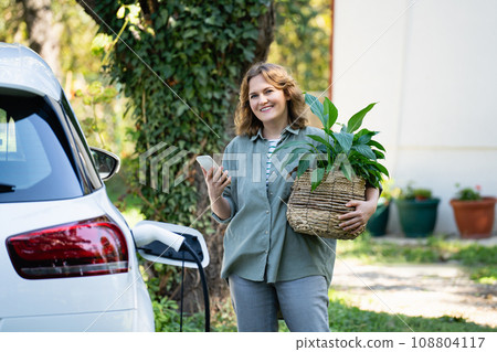 Woman with plant in pot next to a charging electric car in the yard of a country house 108804117