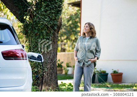 Woman next to a charging electric car in the yard of a country house Woman next to a charging electric car in the yard of a country house 108804121