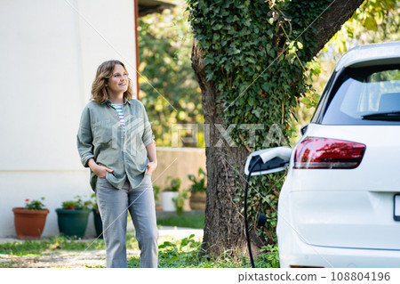 Woman next to a charging electric car in the yard of a country house Woman next to a charging electric car in the yard of a country house 108804196