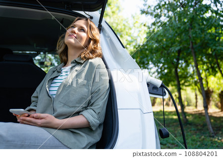 Woman with smartphone sits in an electric car's trunk Woman with smartphone sits in an electric car's trunk 108804201