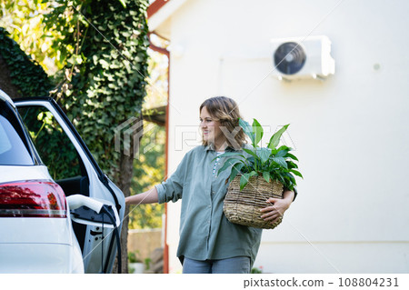 Woman with plant in pot next to a charging electric car in the yard of a country house Woman with plant in pot next to a charging electric car in the yard of a country house 108804231