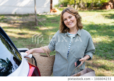 Woman with shopping bag next to a charging electric car in the yard of a country house Woman with shopping bag next to a charging electric car in the yard of a country house 108804233