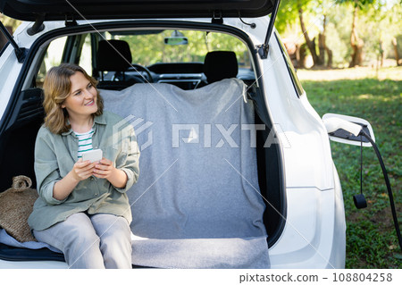 Woman with smartphone sits in an electric car's trunk 108804258