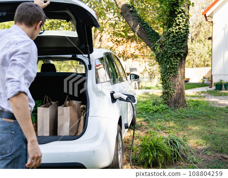 Man with shopping bag next to a charging electric car in the yard of a country house Man with shopping bag next to a charging electric car in the yard of a country house 108804259
