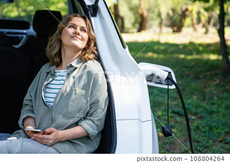 Woman with smartphone sits in an electric car's trunk 108804264
