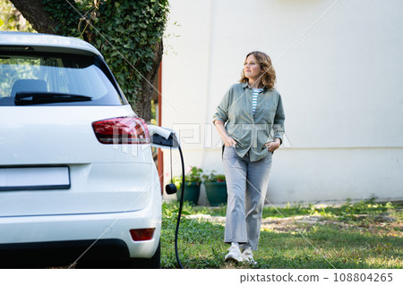 Woman next to a charging electric car in the yard of a country house Woman next to a charging electric car in the yard of a country house 108804265