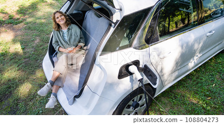 Woman with smartphone sits in an electric car's trunk Woman with smartphone sits in an electric car's trunk 108804273