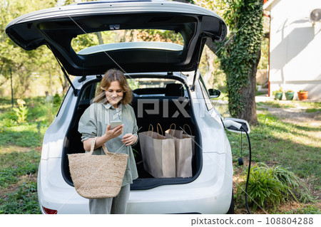 Woman with shopping bag next to a charging electric car in the yard of a country house Woman with shopping bag next to a charging electric car in the yard of a country house 108804288