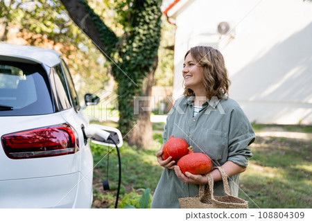 Woman with shopping bag and pumpkins next to a charging electric car in the yard of a country house 108804309