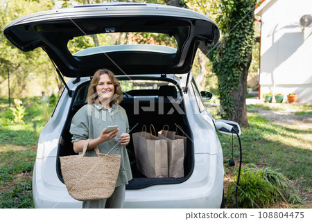 Woman with shopping bag next to a charging electric car in the yard of a country house 108804475