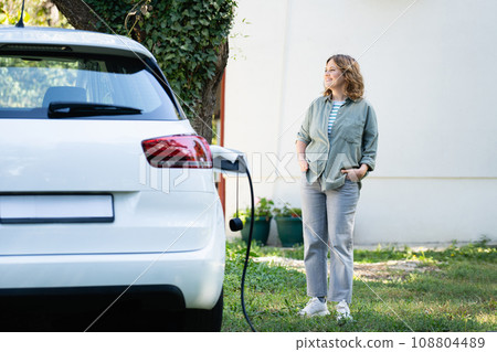 Woman next to a charging electric car in the yard of a country house Woman next to a charging electric car in the yard of a country house 108804489