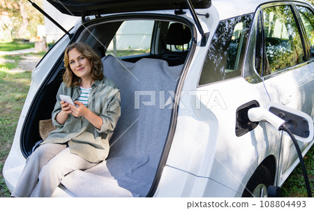 Woman with smartphone sits in an electric car's trunk 108804493