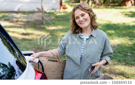 Woman with shopping bag next to a charging electric car in the yard of a country house 108804510