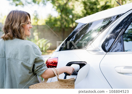 Woman with shopping bag next to a charging electric car in the yard of a country house 108804523