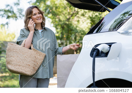 Woman with shopping bag next to a charging electric car in the yard of a country house Woman with shopping bag next to a charging electric car in the yard of a country house 108804527