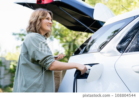 Woman with shopping bag next to a charging electric car in the yard of a country house Woman with shopping bag next to a charging electric car in the yard of a country house 108804544