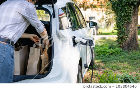 Man with shopping bag next to a charging electric car in the yard of a country house 108804546