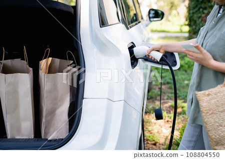 Woman with shopping bag next to a charging electric car in the yard of a country house Woman with shopping bag next to a charging electric car in the yard of a country house 108804550