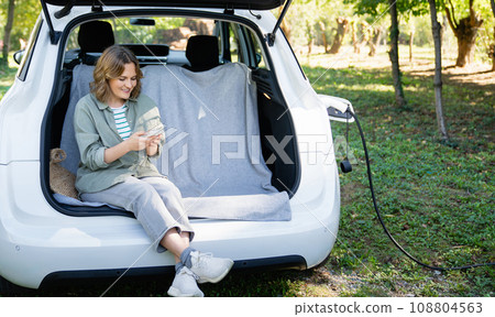 Woman with smartphone sits in an electric car's trunk 108804563