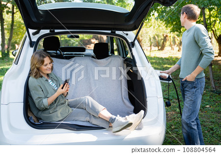 Woman with smartphone sits in an electric car's trunk when the man holds electric plug 108804564