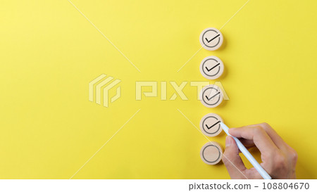 Checklist concept. Person using a black ink pen to correct sign and tick check mark on wooden block with copy space for the checklist on yellow background. Business survey questionnaire, exam process, 108804670