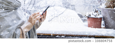 A woman in a warm hat and blanket holds smartphone in her hands. Winter landscape with snowfall in the background A woman in a warm hat and blanket holds smartphone in her hands. Winter landscape with snowfall in the background 108804758