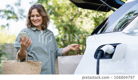 Woman with shopping bag next to a charging electric car in the yard of a country house 108804797