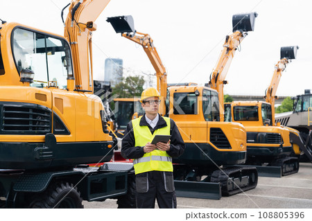 Engineer in a helmet with a digital tablet stands next to construction excavators. 108805396