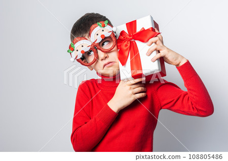 Surprised sad boy Gen Alpha in a red christmas holidays sweater and funny party glasses. Stylish boy with a Christmas gift have joyful on a white background. Generation Alpha Surprised sad boy Gen Alpha in a red christmas holidays sweater and funny party glasses. Stylish boy with a Christmas gift have joyful on a white background. Generation Alpha 108805486