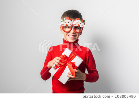 Smiling child boy wearing fun glasses in form of Christmas Santa Claus holding Xmas gift while standing in a white background, dressed in warm red xmas sweater. Generation alpha and gen alpha children 108805490