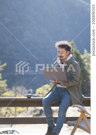 Autumn/winter of a freelance-looking man working remotely with a computer on a wooden deck in the countryside, looking at the camera Autumn/winter of a freelance-looking man working remotely with a computer on a wooden deck in the countryside, looking at the camera 108806503