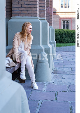 Smiling young woman sits on bench with bouquet of flowers and waits for someone. Red-haired woman outdoors on walk. Vertical frame. Smiling young woman sits on bench with bouquet of flowers and waits for someone. Red-haired woman outdoors on walk. Vertical frame. 108806727
