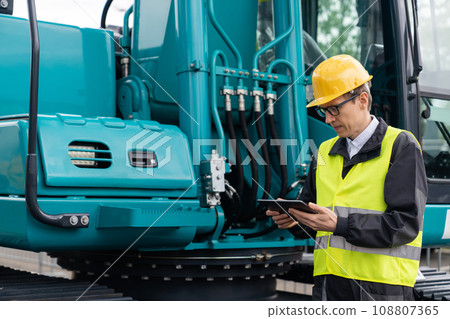 Engineer in a helmet with a digital tablet stands next to construction excavators. Engineer in a helmet with a digital tablet stands next to construction excavators. 108807365