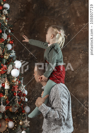 Little girl sits on dads shoulders near beautiful Christmas tree. Portrait of young father and daughter near Christmas tree. Little girl sits on dads shoulders near beautiful Christmas tree. Portrait of young father and daughter near Christmas tree. 108807719