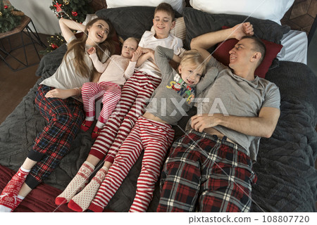 Happy young family wear pajamas and lie on large bed. Mother father and three children. Top view. Christmas or New Year Morning. Holidays Happy young family wear pajamas and lie on large bed. Mother father and three children. Top view. Christmas or New Year Morning. Holidays 108807720