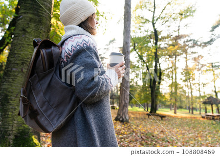 Woman traveler with backpack is holding a cup of hot drink in autumn park.. 108808469