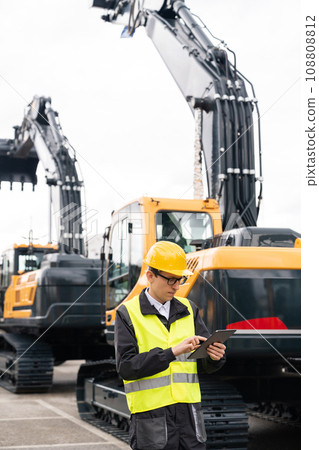 Engineer in a helmet with a digital tablet stands next to construction excavators 108808812