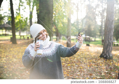 A woman is holding a phone and a cup of hot drink. Woman taking selfie on smartphone 108808827