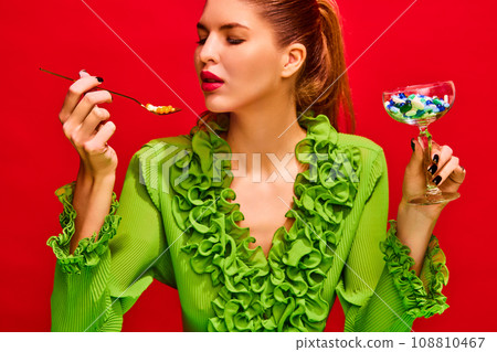 Close up portrait of young pretty lady eating meal, drinking cocktail of tablets, pills and vitamins isolated red background. 108810467