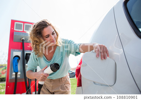 A woman stands at the charging station and holds a plug of the charger 108810901