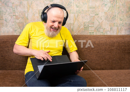 bearded man wearing headphones uses a laptop while relaxing on his home sofa. bearded man wearing headphones uses a laptop while relaxing on his home sofa. 108811117