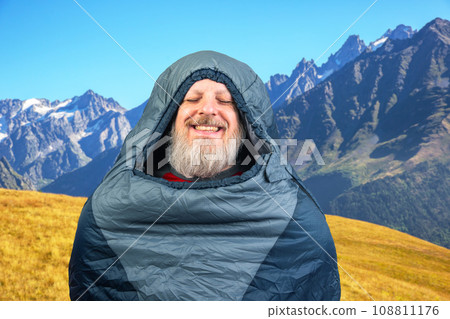 Happy bearded man in a sleeping bag against the backdrop of nature in the mountains. Happy bearded man in a sleeping bag against the backdrop of nature in the mountains. 108811176