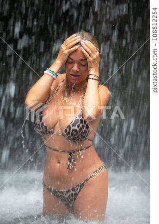 Beautiful young woman relaxes under a waterfall. Sexy girl in a bikini posing near a waterfall in the tropics. A traveler bathes under streams of water flowing from the mountains. 108812254