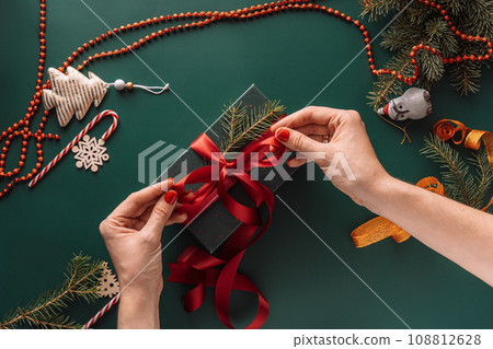 Hands tying red bow on gift on green background with Christmas tree decorations 108812628