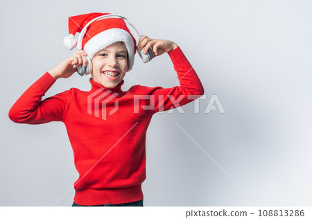 Funny child boy holds listening to Christmas music with Santa Claus hat on white background with copy space. Generation alpha and gen alpha children. Funny child boy holds listening to Christmas music with Santa Claus hat on white background with copy space. Generation alpha and gen alpha children. 108813286