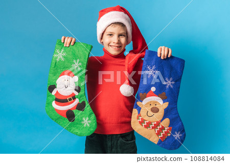Child boy in red sweater and Santa hat with Christmas socks on blue background with space for text Child boy in red sweater and Santa hat with Christmas socks on blue background with space for text 108814084