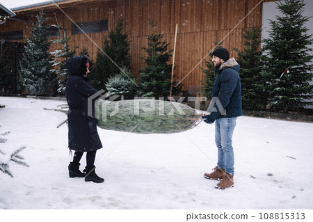 Son with her mom carry Christmas tree to the car. Live conifer from eco store in village. Anticipation of the holiday. Winter mood Son with her mom carry Christmas tree to the car. Live conifer from eco store in village. Anticipation of the holiday. Winter mood 108815313