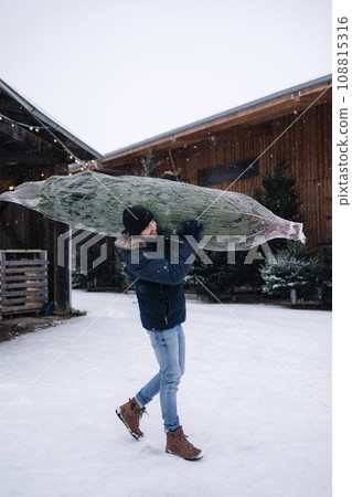 Handsome bearded man holding wrapped Christmas tree on fair. Happy male bring fir tree. Handsome bearded man holding wrapped Christmas tree on fair. Happy male bring fir tree. 108815316